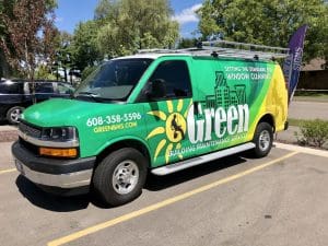 A green van featuring Fleet Wraps and Graphics for “Green Building Maintenance Services” is parked in a lot. The company’s contact info, cityscape, sun design, and window cleaning graphics are proudly displayed on the side.