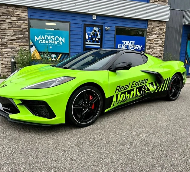 A bright green sports car with black rims and Real Estate lettering—showcasing creative vehicle wraps—is parked in front of the Madison Graphics Company building with stone and blue siding, featuring signs for graphics and tinting services.